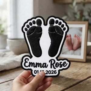 May include: A white fabric patch featuring black embroidered baby footprints at the top. Below the footprints, the name Emma Rose is embroidered in black script, followed by the date 01.15.2026 in black text, all outlined in black.