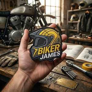 May include: A black, yellow, and white embroidered patch shaped like a motorcycle helmet with the text BIKER JAMES in bold lettering. The patch features a detailed visor and shell design, showcasing clean stitching and a high-contrast color scheme.