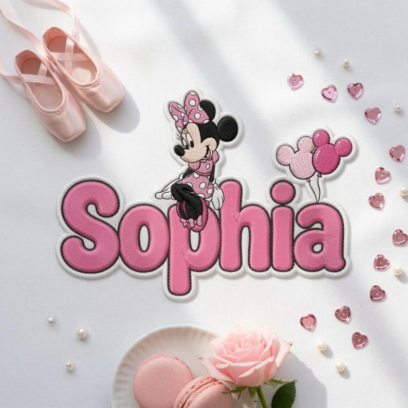 Op de afbeelding: Een gepersonaliseerde geborduurde patch met de naam Sophia in roze letters, geaccentueerd door een Minnie Mouse-figuur met een gestippelde strik en twee Mickey-vormige ballonnen, omringd door roze hartvormige lovertjes en witte parels.