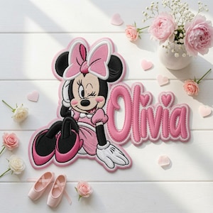 Może przedstawiać: Naszywka z tkaniny przedstawiająca puszczającą oko Myszkę Minnie w różowej sukience i kokardzie, obok imienia Olivia wyhaftowanego różową kursywą z akcentami w kształcie serc. Wzór posiada białe obwódki i szczegółowe przeszycia.