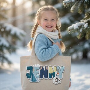 Puede incluir: Un bolso de tela beige con el nombre JENNY en letras grandes, coloridas y estilo bloque. La letra J incorpora un gr&aacute;fico de un personaje rubio con vestido azul, y la letra Y presenta un peque&ntilde;o gr&aacute;fico de un mu&ntilde;eco de nieve blanco.