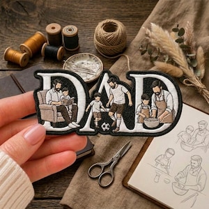 May include: A black and white embroidered patch featuring the word DAD, with illustrations of a person reading to a child, a person playing soccer with a child, and a person cooking with a child, all rendered in detailed black thread on white fabric.