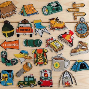 May include: A collection of embroidered patches with a camping theme. Patches include a tent, compass, hiking sign, backpack, lantern, and a vehicle with the text "STAY WILD". The color palette includes greens, browns, oranges, and blues.