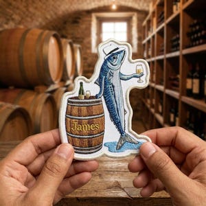 May include: An embroidered patch featuring a blue fish wearing a white fedora, holding a wine glass, and leaning against a brown wooden barrel. The barrel has the name James written on it in yellow text, with a small bottle and glass on top.