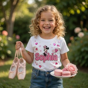 Op de afbeelding: Een wit T-shirt met korte mouwen voorzien van een Minnie Mouse-afbeelding, roze hartvormige accenten, parelachtige versieringen en de naam Sophia in roze letters. Het shirt wordt gecombineerd met een blauwe spijkerbroek en lichtroze balletschoenen.