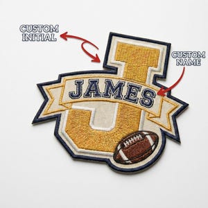 May include: Embroidered patch with a gold "J" initial, a banner reading "JAMES", and a football. The patch has a navy blue border and is designed for custom names and initials. The patch is a sports-themed item.