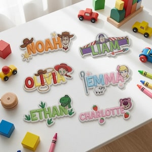 May include: Six embroidered name patches featuring Toy Story characters. The patches display the names Noah, Liam, Olivia, Emma, Ethan, and Charlotte, each decorated with themed icons like cowboy hats, space ranger wings, lassos, sheep, dinosaurs, and strawberries.