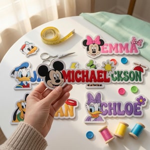 Peut inclure: Une collection d'écussons en tissu brodé représentant des personnages Disney tels que Mickey, Minnie, Donald, Daisy et Pluto. Les écussons affichent des prénoms comme Emma, Michael et Chloe avec des lettres colorées, épaisses et brodées.