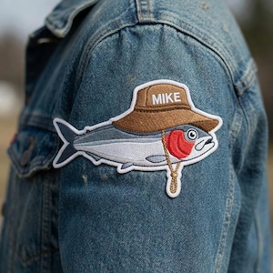May include: Embroidered patch featuring a cartoon fish wearing a brown hat with the name "MIKE" on it. The fish has a red gill and a gold-colored string. The patch is sewn onto a blue denim jacket.
