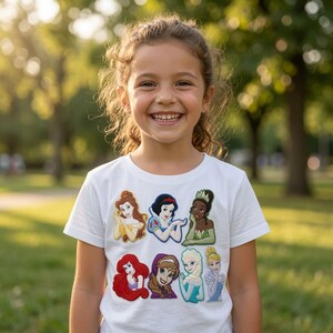 May include: A white short-sleeved t-shirt featuring a graphic print of seven animated female characters arranged in two rows. The characters include figures with brown, black, red, blonde, and white hair wearing colorful dresses in yellow, blue, green, and purple.