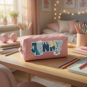 Puede incluir: Un estuche rectangular de color rosa con el nombre JENNY en letras grandes y coloridas. La letra J incluye la ilustraci&oacute;n de un personaje con vestido azul, y la letra Y presenta un peque&ntilde;o mu&ntilde;eco de nieve. Tiene un tirador de cremallera en forma de coraz&oacute;n.