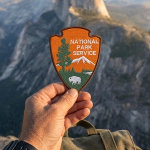 May include: An embroidered National Park Service patch in the shape of an arrowhead with a brown border. The design features an orange sky, a green tree, a white mountain range, and a white bison, with the text NATIONAL PARK SERVICE in white.