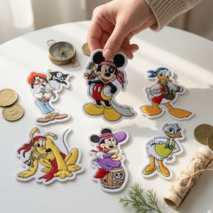 May include: A collection of six embroidered fabric patches featuring cartoon characters dressed as pirates, including Mickey Mouse, Minnie Mouse, Donald Duck, Daisy Duck, Goofy, and Pluto, arranged with gold coins and a rolled map.