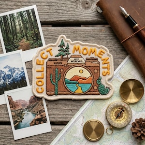May include: Embroidered patch with a camera design and the words "Collect Moments" and "Keep Nature Wild." The patch features a desert landscape with a cactus and a lake. Other items include a compass, map, and photos on a wooden surface.