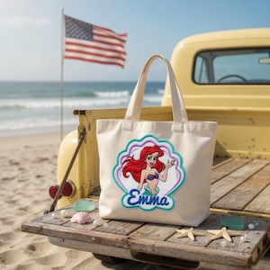 May include: A cream-colored canvas tote bag featuring an embroidered patch of a red-haired mermaid character inside a scalloped shell shape. The name Emma is embroidered in blue script below the character. The bag has two sturdy top handles.