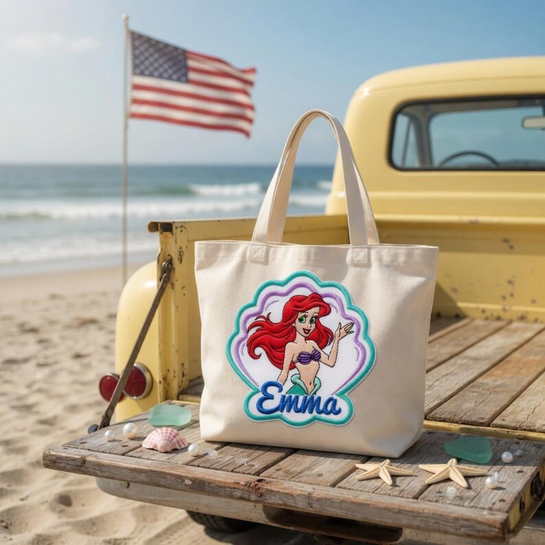 May include: A cream-colored canvas tote bag featuring an embroidered patch of a red-haired mermaid character inside a scalloped shell shape. The name Emma is embroidered in blue script below the character. The bag has two sturdy top handles.