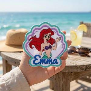 May include: A fabric patch shaped like a seashell featuring an embroidered illustration of a red-haired mermaid with a purple seashell top and a green tail. The name Emma is embroidered in blue script at the bottom, outlined in teal and purple.