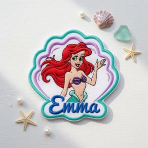 May include: A shell-shaped embroidered patch featuring a cartoon mermaid with long red hair, a purple seashell top, and a green tail. The name Emma is embroidered in blue script at the bottom. The patch has a white background with teal and purple borders.