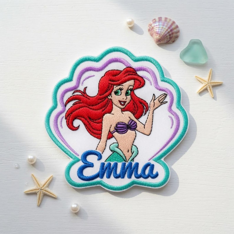 May include: A shell-shaped embroidered patch featuring a cartoon mermaid with long red hair, a purple seashell top, and a green tail. The name Emma is embroidered in blue script at the bottom. The patch has a white background with teal and purple borders.