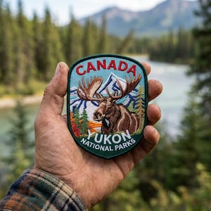 May include: An embroidered patch with a dark green border featuring a brown moose, blue mountains, and green pine trees. The text CANADA is arched at the top in red, and YUKON NATIONAL PARKS is printed in white at the bottom of the shield-shaped design.