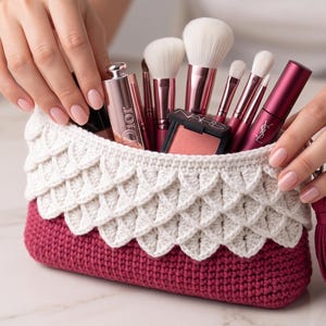 May include: A handmade crochet makeup bag in burgundy and white, filled with makeup brushes, blush, and other cosmetics. The bag has a scalloped design and a matching tassel. The bag is approximately 8 inches long.