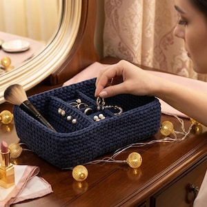 May include: A navy blue, crocheted jewelry organizer with multiple compartments, holding rings, earrings, and bracelets. A makeup brush and perfume bottle are visible on the wooden surface. Decorative lights and a mirror are in the background.