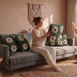 Könnte beinhalten: Ein gemütliches Wohnzimmer mit einem grauen Sofa, das mit grünen Häkelkissen geschmückt ist. Jedes Kissen zeigt ein großes, gänseblümchenartiges Blumendesign mit cremefarbenen und türkisfarbenen Akzenten. Der Raum ist in warmen, neutralen Tönen gehalten.