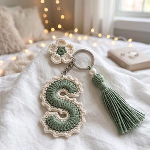 May include: A handmade crochet keychain featuring the letter "S" in sage green with a cream-colored border. The keychain includes a matching flower, a green tassel, and a pearl accent. The design is attached to a silver keyring.