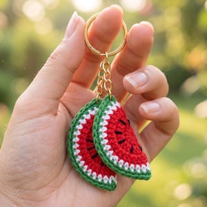 May include: A handmade keychain featuring two crochet watermelon slices. The watermelon slices are red with black seed details, green rinds, and white accents. The keychain has a gold-coloured ring and chain.