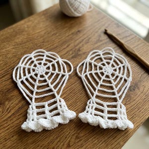 May include: A pair of white crocheted fingerless gloves featuring a spiderweb pattern that extends from the wrist to the knuckles, finished with a ruffled edge at the wrist opening, crafted with fine white yarn and a wooden crochet hook.
