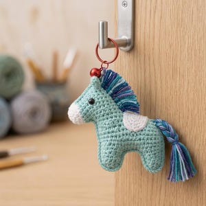 May include: A crocheted horse keychain in shades of teal, white, and blue, hanging from a metal hook. The handmade horse has a braided tail and a small bell. Yarn and crochet hooks are in the background.
