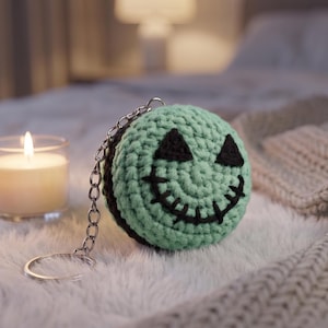 May include: A crocheted keychain featuring a green and black Jack Skellington design. The round keychain has a smiling face with triangular eyes and a stitched mouth. A lit candle in a glass holder sits nearby.