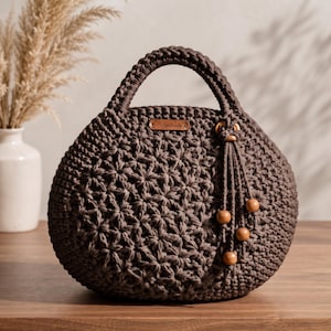 May include: A round, dark brown crochet handbag featuring a textured star-like pattern on the front. The bag has a sturdy integrated handle, a small rectangular wooden tag labeled "Handmade," and a decorative tassel with four smooth wooden beads.