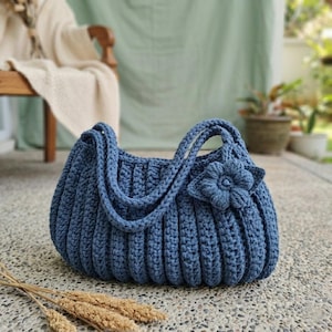 May include: A blue crochet handbag with a flower accent. The bag features a ribbed design and two handles. The bag is approximately 12 inches wide and 8 inches tall.