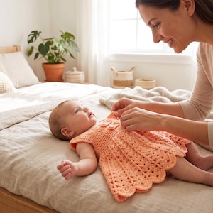 May include: A baby wearing a peach-colored, crocheted dress with a scalloped hem lies on a bed. A woman is adjusting the dress. The room has a neutral color palette with a wooden bed frame and a potted plant.