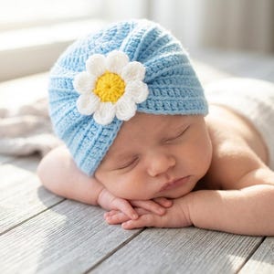 May include: A sleeping baby wearing a light blue crochet hat with a white and yellow daisy. The baby is lying on a wooden surface, with its hands gently supporting its head. The hat is the focal point, showcasing the handmade details.