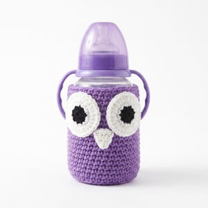 May include: A baby bottle with a purple plastic cap and two side handles, encased in a purple crocheted cozy. The cozy features a large owl face design with two white circular eyes, black pupils, and a small white triangular beak centered.