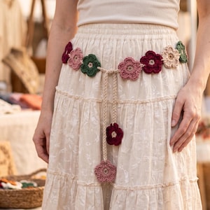 Puede incluir: Una falda de color crema con niveles y un cinturón de flores de ganchillo. El cinturón consta de flores interconectadas en tonos rojo oscuro, verde bosque y rosa claro. Dos cordones trenzados cuelgan del centro, terminando en una flor.