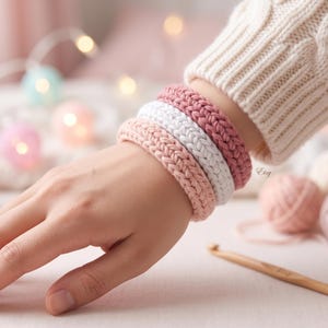 以下が含まれることがあります： ピンクと白の色合いの手作りの編みブレスレットが4つ、手首に重ねられています。ブレスレットは編み込みのような外観です。クリーム色のセーターを着ています。背景には毛糸玉と編み棒があります。