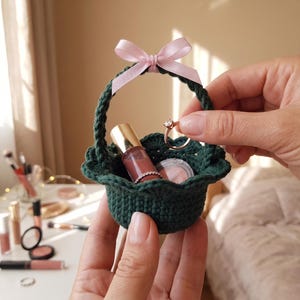May include: A small, dark green crocheted basket with a pink ribbon bow on the handle. The basket contains makeup items, including lip gloss and a compact. A ring is held above the basket.