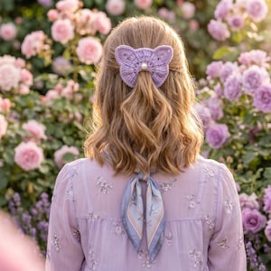 May include: A woman's back is shown, with a light purple crochet butterfly hair clip with a pearl center. She wears a light purple floral blouse and a blue and white scarf. The background is filled with pink and purple roses.