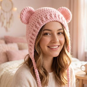 May include: A smiling person wearing a light pink crocheted hat with bear ears. The hat has long ear flaps. The background includes a bed with pink pillows and a dream catcher.