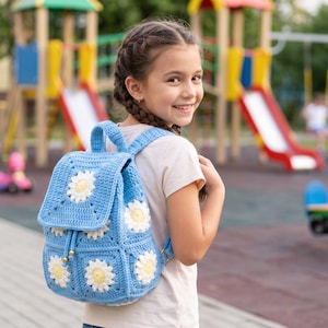 May include: A light blue crochet backpack featuring a pattern of white and yellow daisy motifs. The bag has a top handle, adjustable shoulder straps, and a drawstring closure with metallic toggles, constructed from yarn in a square stitch design.