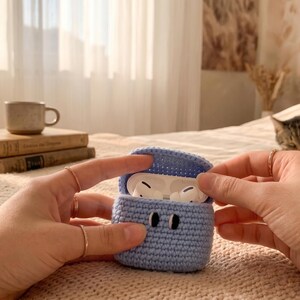 May include: A pale blue, crocheted case for wireless earbuds, with a matching lid. The case is open, revealing white earbuds inside. The case has two black stitched eyes. The background includes a bed, books, and a cat.