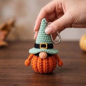 May include: A handmade crochet gnome keychain with a pumpkin-shaped body and a witch hat. The gnome has an orange body, a green hat with a black band and gold buckle, and a beige nose. The keychain is held by a silver clasp.