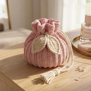 May include: A pink crocheted drawstring pouch with beige leaf accents. The pouch has a tassel and drawstring closure. The pouch is on a wooden surface with beauty products and jewelry.