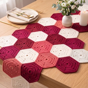 May include: A handmade crochet table runner in a honeycomb pattern. The runner features a gradient of colors, including white, light pink, and deep red. The runner is displayed on a wooden table, alongside a plate, silverware, and a vase of flowers.