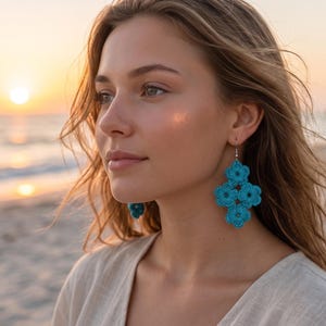 May include: A woman wearing turquoise crochet earrings with a floral design. The earrings are a vibrant blue color and dangle from silver hooks. The background shows a beach scene with the sun setting over the ocean.