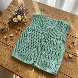 May include: A sleeveless knit vest in a soft sage green color, featuring a textured diamond pattern on the front panels and a ribbed neckline. The garment is secured with three small, round, white pearl-like buttons down the center front.