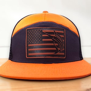 Puede incluir: Una gorra de béisbol de color naranja brillante y azul marino oscuro con un parche rectangular en el panel frontal. El parche muestra un diseño estilizado de la bandera estadounidense con la silueta de un pez marlín en las franjas.